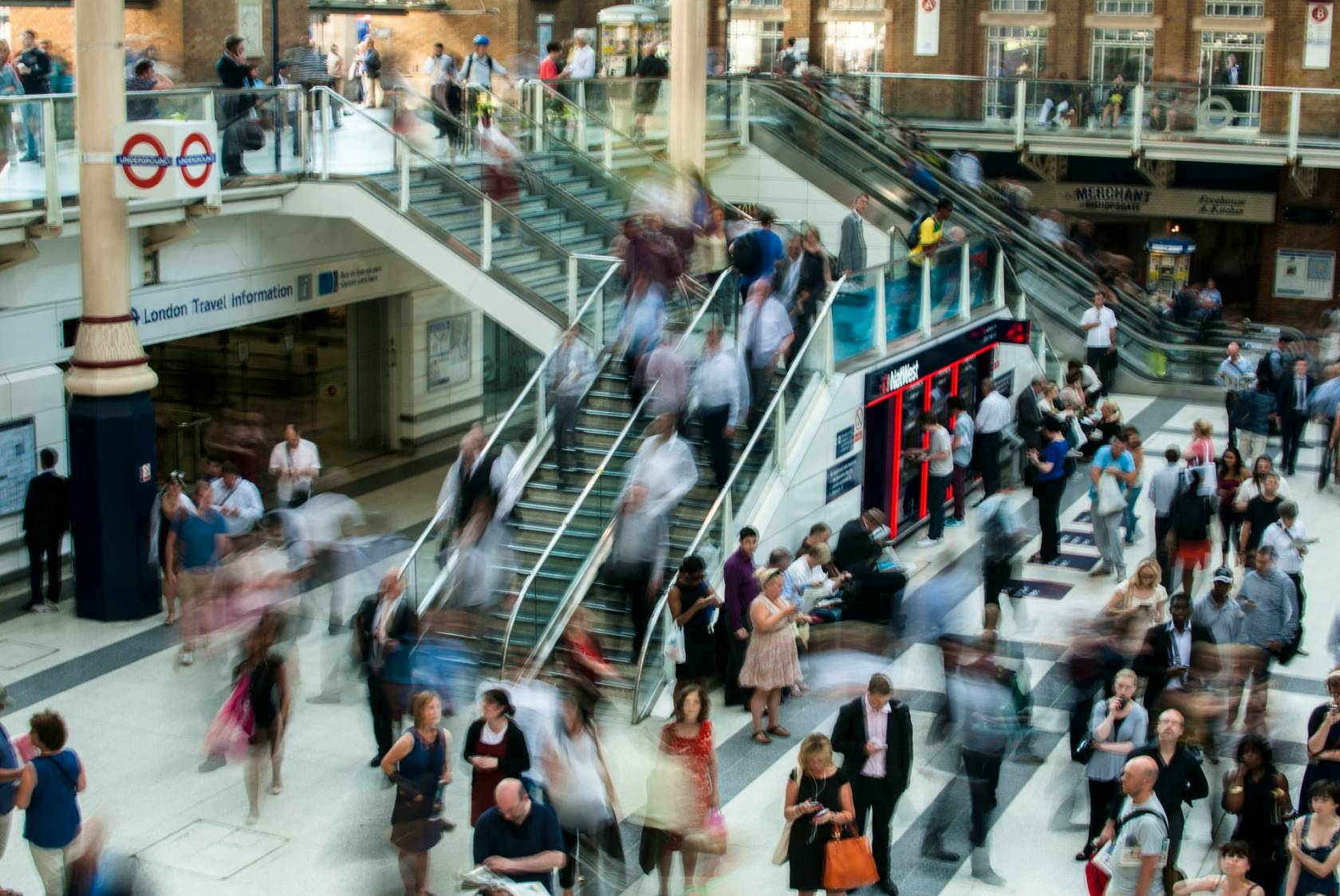 People in jeans walking in a crowded public space in London, with a sign showing travel information and text that reads "00 London Travel information 42322 MERCHANT WISHOPSGATE Preghouse & Allia."